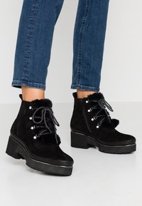 Stivaletti neri in suede con fodera in pelliccia sintetica, caratterizzati da accenti metallici decorativi e suole a zeppa testurizzate. Chiusura con lacci e cerniera laterale.