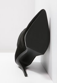 Scarpa nera in suede con tacco alto, punta affusolata e tacco sottile e appuntito. La suola è liscia con una leggera texture. Posizionata su uno sfondo bianco.