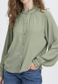 Woman wearing light green blouse with ruffled collar and cuffs, buttoned front, and blue jeans, hand in pocket, against plain background.