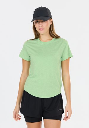 Kvinde iført en lysegrøn kortærmet skjorte, sorte lagdelte shorts og en sort baseballcap står mod en ensartet baggrund.