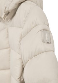Cazadora de plumas beige con textura acolchada, que incluye una capucha y un parche de logo de goma en la manga. Material suave y ligero.