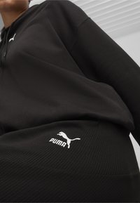 Svart hoodie och ribbade svarta byxor med en vit Puma-logotyp. Materialet verkar mjukt; designen är minimalistisk med en avslappnad passform.