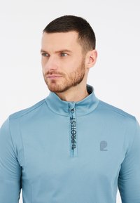 Svetlai mėlyna ilgos rankovės megztinis su pusiau užtrauktuku, turintis subtilų logotipą ir kontrastinėmis siūlėmis ant pečių. Lygus paviršius.