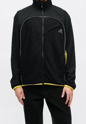 Personne portant une veste zippée noire Adidas avec bordure jaune et une poche poitrine zippée, debout les bras détendus le long du corps.