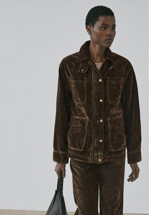 Hnědá manšestrová bunda se čtyřmi kapsami, límečkem a zapínáním na knoflíky. Model drží černou kabelku a má na sobě odpovídající hnědé kalhoty.