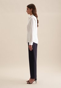 Witte blouse met lange mouwen, gecombineerd met marineblauwe pantalon en zwarte hoge hakken, tentoongesteld tegen een neutrale achtergrond.