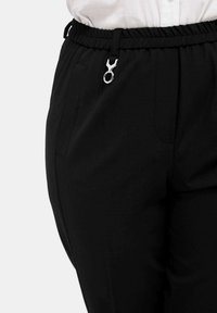 Zwarte elastische broek met een zilveren hardwareaccent. Kenmerken zijn onder andere zijzakken en een gladde, lichte stoftextuur.