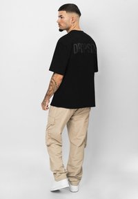Sort oversized t-shirt med "DRIPSIZE" i en tonet tryk på ryggen; parret med lyse beige cargo bukser og hvide sneakers.