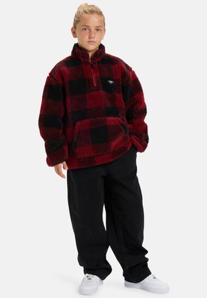 Pull en polaire à carreaux rouge et noir avec un col montant, poche avant et jean noir. Porté avec des baskets blanches.