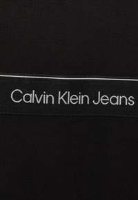 Černá tkanina s výrazným, kontrastním bílým logem s nápisem "Calvin Klein Jeans" nahoře, s hladkou strukturou a plochým designem.