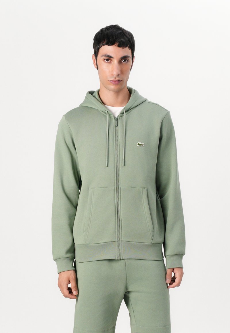 Sudadera con cremallera de color verde claro, con capucha ajustable, bolsillos tipo canguro, puños acanalados y un pequeño logo en el pecho. Fabricada con un tejido suave.