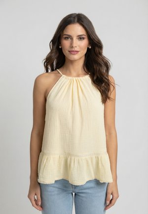 Femme aux longs cheveux bruns portant des boucles d'oreilles en perles et un haut dos nu sans manches jaune clair à ourlet volanté, associée à un jean bleu clair.