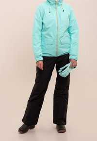 Veste imperméable vert menthe avec fermeture éclair à l'avant, deux poches, assortie à un pantalon de neige noir et des gants vert menthe. Chaussures noires visibles.