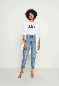 Woman with curly hair wearing white open shirt over white GAP logo tee, high-waist blue jeans, and white heeled sandals standing against white wall.