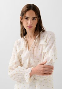 Blouse blanche avec des manches à volants et de subtils motifs carrés dorés. Dotée d'un décolleté en V et de poignets froncés. Tissu léger et texturé.