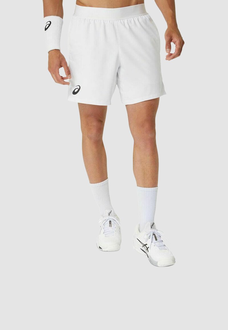 Hombre vistiendo pantalones cortos deportivos blancos de Asics, zapatos deportivos blancos, calcetines blancos y una muñequera blanca con el logo de Asics sobre un fondo liso.
