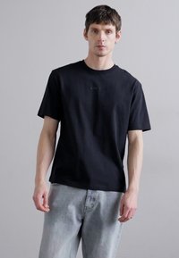 Tricou scurt negru cu mânecă scurtă, guler rotund și text brodat subtil pe piept, purtat cu blugi din denim gri deschis.