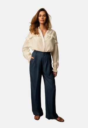 Blusa bianca in lino con dettagli a ruches, apertura con bottoni davanti, abbinata a pantaloni a gamba larga blu navy. Modella che indossa sandali piatti marroni.