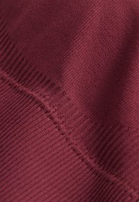 Burgundinpunainen neulottu kangas, jossa on teksturoitu kuviointi, vaakasuoria ja pystysuoria ribbineuloksia sekä näkyvät sauman yksityiskohdat.