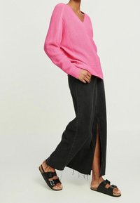 Rosa gerippter Strick-V-Ausschnitt-Pullover, schwarze weit geschnittene Denim-Rock mit einem Schlitz vorne und schwarze Slipper mit doppelten Schnallen.