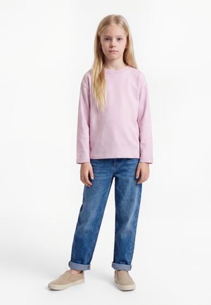 Joven chica rubia de pie con las manos relajadas a los lados, vistiendo una camiseta de manga larga rosa claro, vaqueros azules remangados y zapatos beige sin cordones.