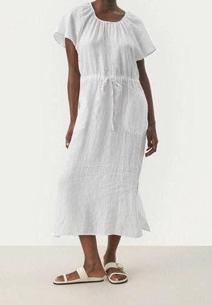 Kobieta ubrana w białą, krótkorękawową sukienkę sięgającą kostek z sznurkiem w talii oraz kremowe sandały z klamrą i paskiem na palce.