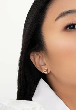 Orecchini a forma di cuore in oro esposti all'orecchio di un modello, caratterizzati da un design vuoto e una superficie liscia. Lo sfondo è bianco semplice, migliorando la visibilità.