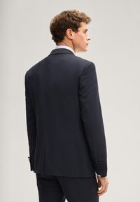 Giacca da completo sartoriale blu navy con colletto a lancia, vestibilità slim e polsini con bottoni. Tessuto liscio, leggermente lucido, senza decorazioni visibili.