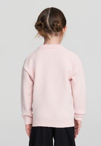 Bambina con capelli castani raccolti in due bun che indossa un maglione rosa chiaro e pantaloni neri, di spalle su uno sfondo grigio uniforme.