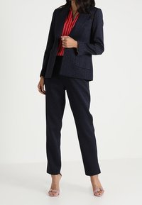 Granatowy garnitur w wzory, składający się z dopasowanej marynarki z kieszeniami oraz spodni o prostych nogawkach, zestawiony z czerwoną bluzką w paski i różowymi szpilkami.