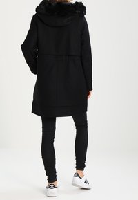 Persona che indossa un cappotto nero con cappuccio e bordo in pelliccia, pantaloni skinny neri e sneakers bianche Adidas, in piedi di spalle su uno sfondo neutro.