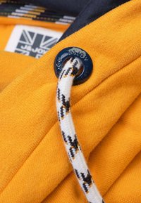 Gros plan sur un tissu jaune avec une bordure bleu marine, comportant un œillet rond bleu marine et un cordon de serrage texturé avec un motif noir et orange.