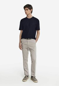 Marinblå kortärmad t-shirt, beige byxor, grå sneakers med svarta detaljer. Modellen står framför en neutral bakgrund, med händerna längs sidorna.