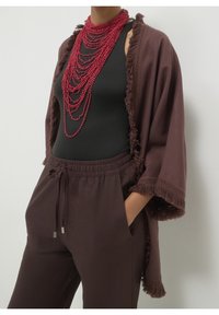 Personne portant des colliers de perles rouges superposés, un haut noir sans manches, un pantalon ample marron foncé avec cordon de serrage et un châle marron à franges, mains dans les poches.