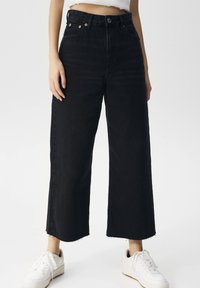 Svarta vida jeans med hög midja, kopparknappdetaljer och fransad nederkant. Matchas med vita sneakers.