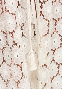 Tissu en dentelle blanche avec des motifs floraux et un pompon tressé suspendu au centre.