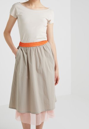Frau trägt ein beige kurzärmeliges Oberteil und einen beigen Rock mit einem orangen elastischen Bund und pinkem Netzsaum, steht mit einer Hand in der Tasche.