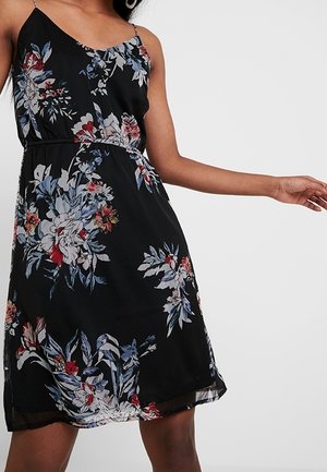 Femme portant une robe noire sans manches avec des motifs floraux bleus, rouges et blancs sur un fond blanc uni.