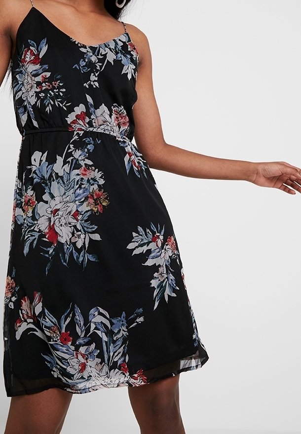 Femme portant une robe noire sans manches avec des motifs floraux bleus, rouges et blancs sur un fond blanc uni.