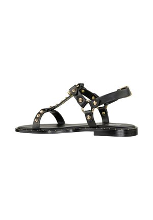 Sandalias de cuero negro con suela plana, que presentan acentos de tachuelas doradas y un diseño de tira en forma de T. Con la parte trasera abierta, son minimalistas y cuentan con un cierre de hebilla.