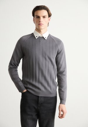 Szary prążkowany sweter z długimi rękawami i okrągłym dekoltem, nałożony na białą koszulę z kołnierzykiem. Całość uzupełniają ciemnoniebieskie dżinsy.