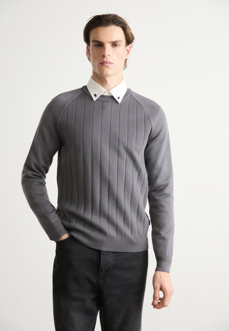 Grauer gerippter Pullover mit langen Ärmeln und rundem Ausschnitt, über ein weißes Hemd mit Kragen getragen. Dunkle Jeans vervollständigen das Outfit.