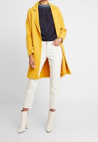 Cappotto giallo, maglia blu navy con colletto a righe, jeans bianchi a vita alta e stivaletti beige. Tessuto morbido, vestibilità su misura e linee pulite.