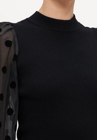 Top nero a coste con collo alto; presenta maniche a sbuffo trasparenti decorate con pois neri.