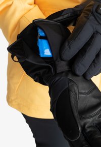 Gants noirs avec paumes en cuir et partie supérieure en tissu, dotés d'une poche zippée contenant un objet bleu. Main positionnée contre une veste jaune.