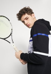 Černobílá mikina s kapucí a modrými pruhy, držící tenisovou raketu se žlutou a černou síťkou, na jednobodovém pozadí.