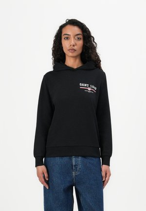 Felpa nera con cappuccio caratterizzata da un logo frontale "GANT 1949" in bianco e rosso. Realizzata in materiale morbido con polsini e orlo a coste. Abbinata a jeans blu.