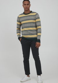 Pulover tricotat cu un model multicolor, cu dungi galbene, verzi și albe; guler și mansete ribbed; purtat cu pantaloni închiși la culoare și adidași albi.