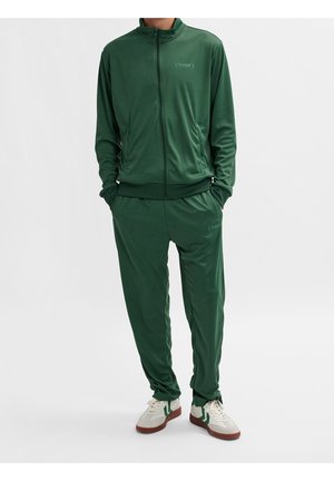 Personne portant un survêtement vert foncé zippé, les mains dans les poches, et des baskets blanches avec des bandes vertes et des semelles marron.