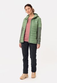 Lichtgroene pufferjack met capuchon, gecombineerd met een roze overhemd, zwarte broek en beige laarzen. Toont een slim-fit ontwerp met een gequilt textuur.